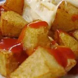 Primer plano de patatas fritas doradas cortadas en cuadrados con un chorrito de salsa roja encima y una cucharada de salsa blanca.