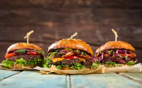 Tres hamburguesas gourmet con distintos ingredientes y brochetas de madera, presentadas en papel pergamino sobre una superficie de madera azul desgastada.