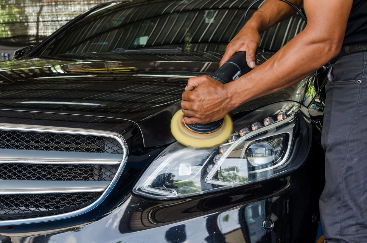 Persona puliendo el faro y el capó de un coche negro con una máquina pulidora.