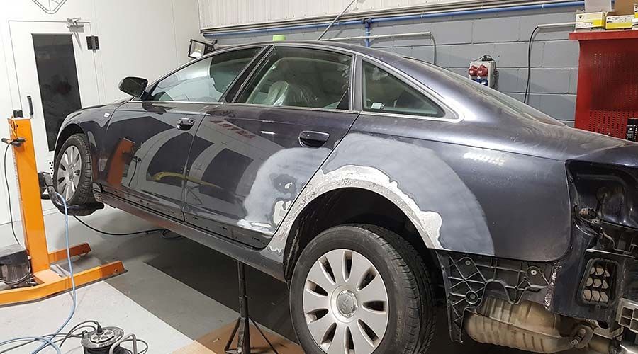 Automóvil gris oscuro en un taller de reparación; el guardabarros trasero y el panel de la puerta muestran trabajos de reparación e imprimación, montado en un elevador.
