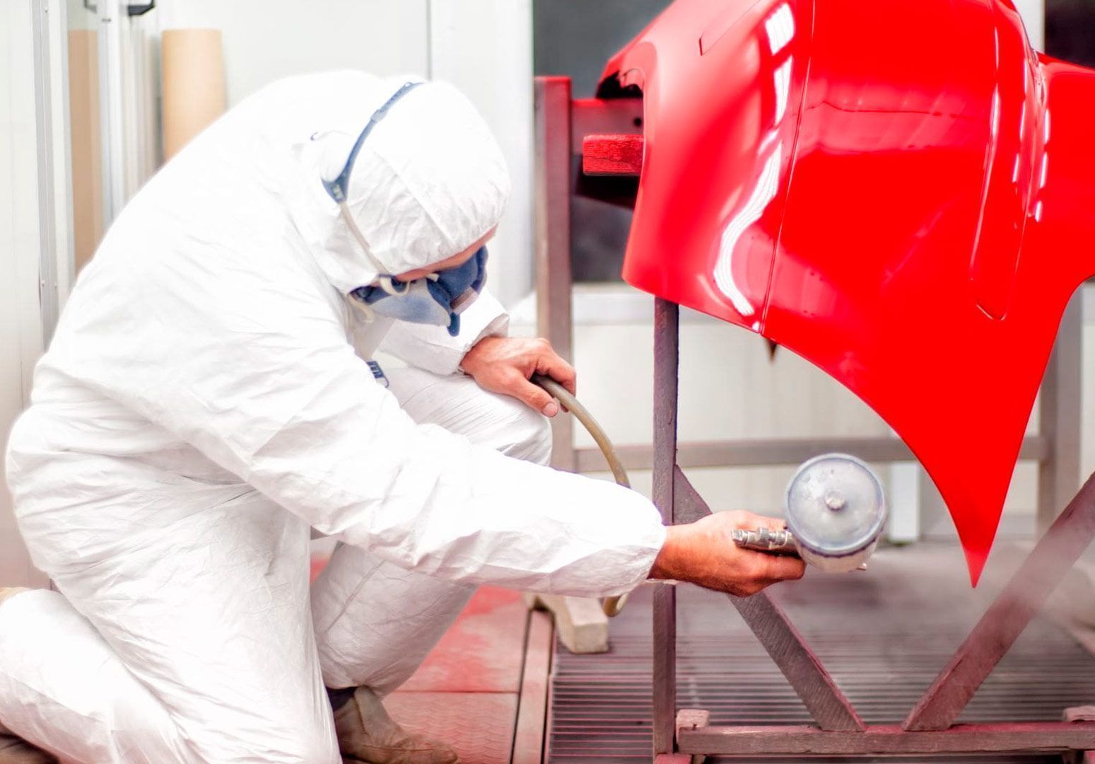 Persona con traje de protección pintando con aerosol el parachoques de un coche rojo en un taller.