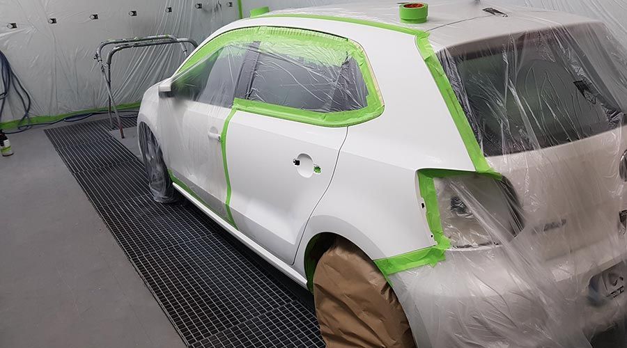 Coche blanco en cabina de pintura, protegido con cinta verde y láminas de plástico, listo para pintar.