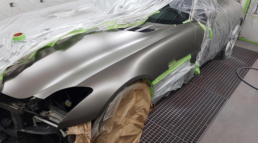 Un coche siendo pintado en un garaje; el coche está cubierto de plástico con secciones selladas con cinta adhesiva, mostrando un acabado gris mate.