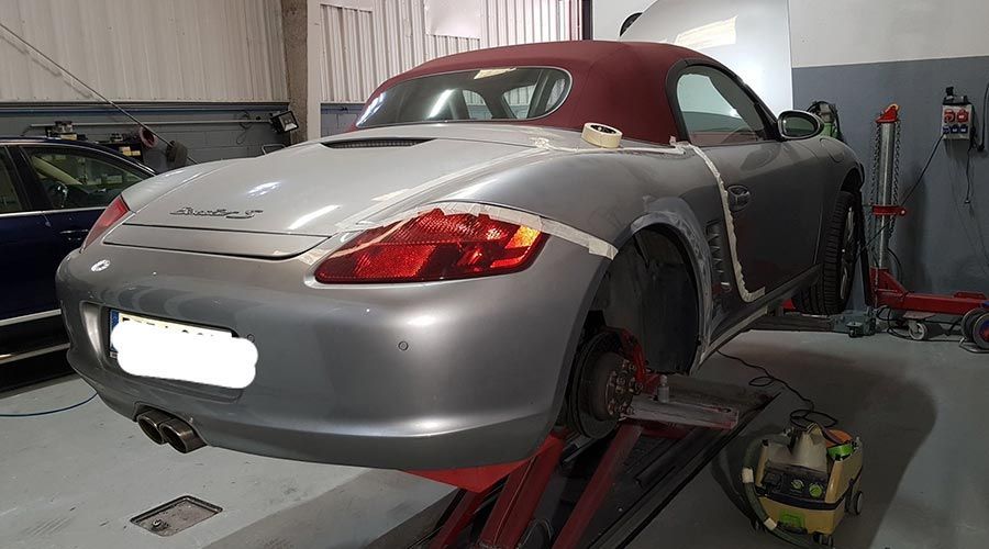 Un Porsche Boxster descapotable plateado con capota roja se eleva en un elevador en un garaje. Se está reparando el panel trasero.