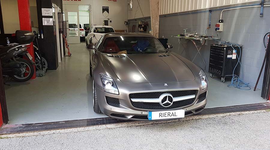 Un Mercedes-Benz SLS AMG plateado estacionado dentro de un garaje, con motocicletas visibles a la izquierda y un automóvil blanco al fondo.