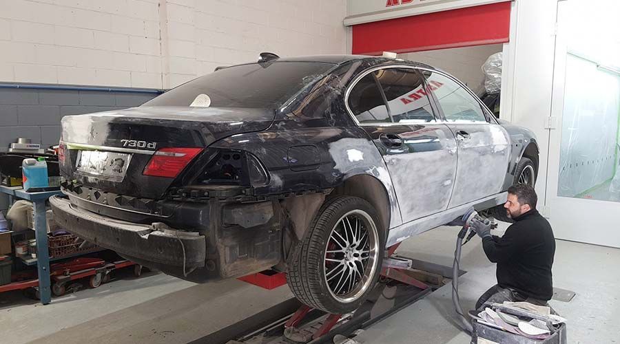 Un mecánico lija un coche negro subido a una plataforma elevadora en un taller. La carrocería tiene zonas con imprimación.