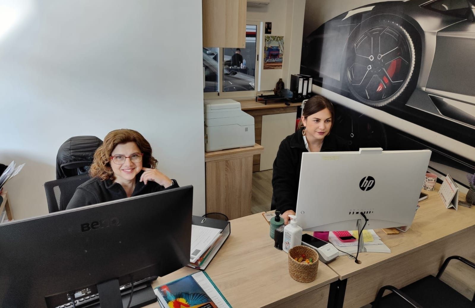 Dos mujeres sentadas en un escritorio de oficina, ambas frente a pantallas de ordenador. La mujer de la izquierda sonríe; la otra está concentrada en su pantalla.