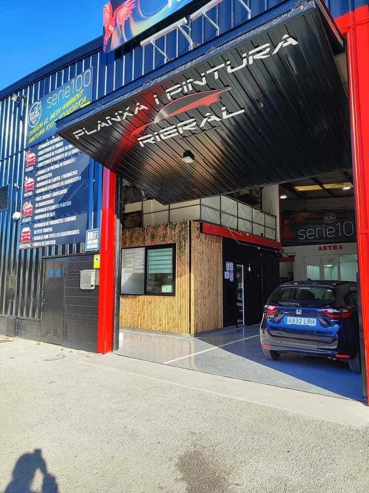 Un taller mecánico con un toldo negro y rojo. Un coche azul está aparcado dentro. El nombre del taller es "PLANXA I PINTURA RIERAL".