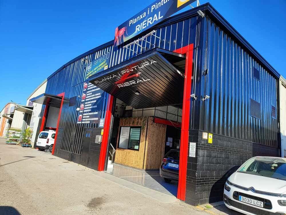Un taller de carrocería negro y rojo con una puerta parcialmente abierta, que muestra un vehículo en su interior, bajo un cielo azul brillante.