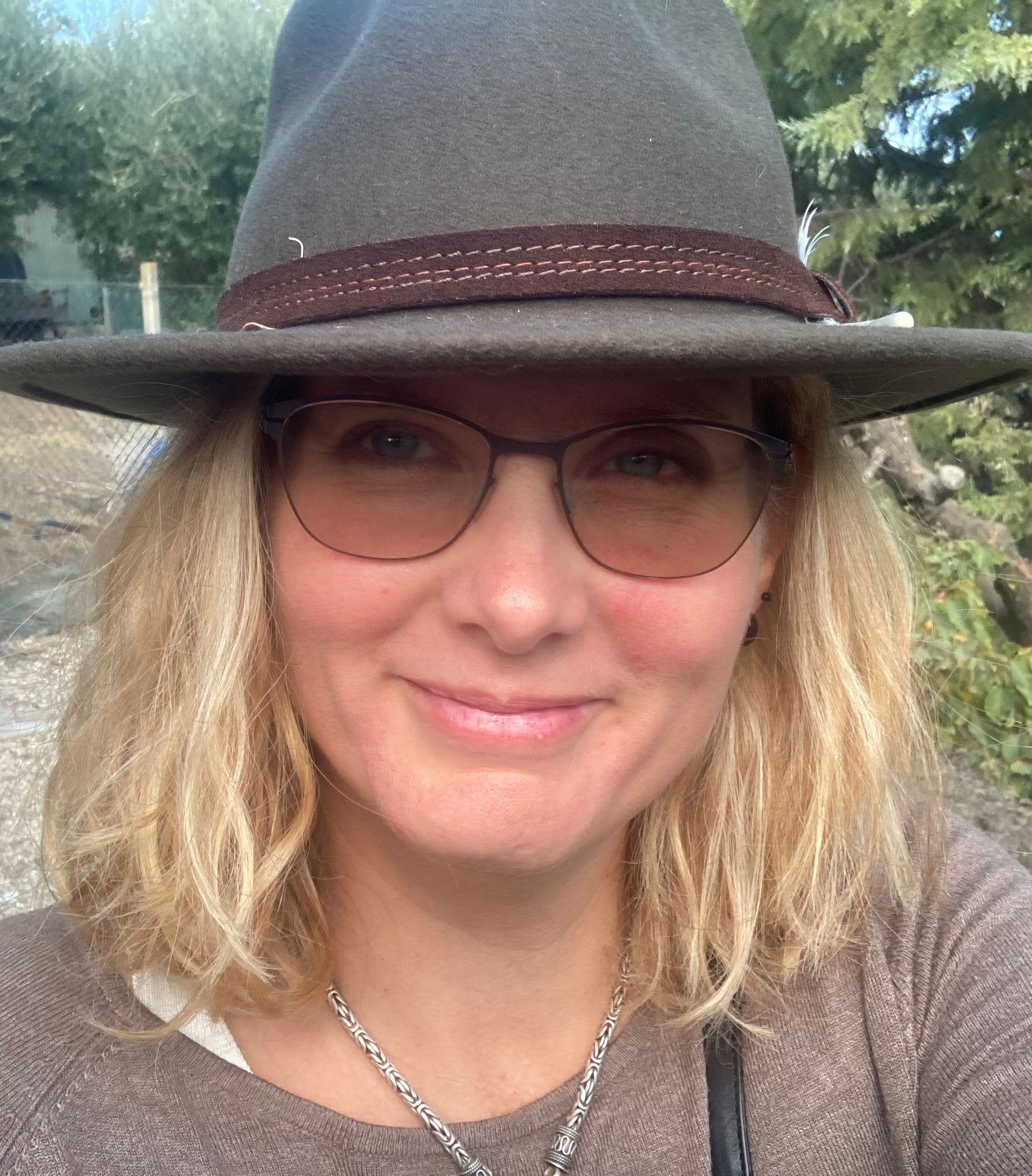 Eine Frau mit kastanienbraunem Haar und blauen Augen lächelt; sie trägt ein grünes, besticktes Oberteil und hat die Hand im Haar.