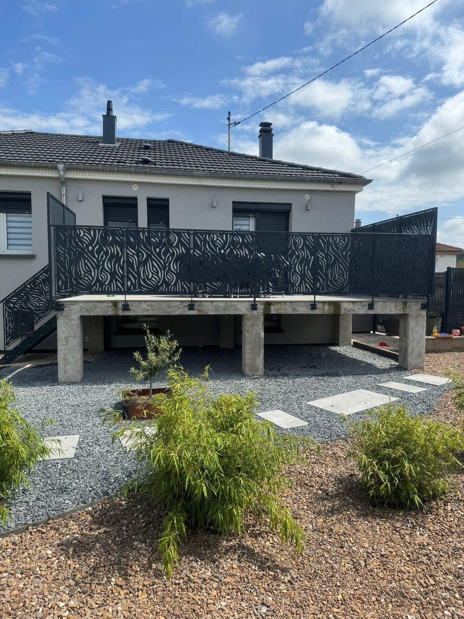 Arrière d'une maison gris clair avec une terrasse décorative en métal noir et un aménagement paysager.