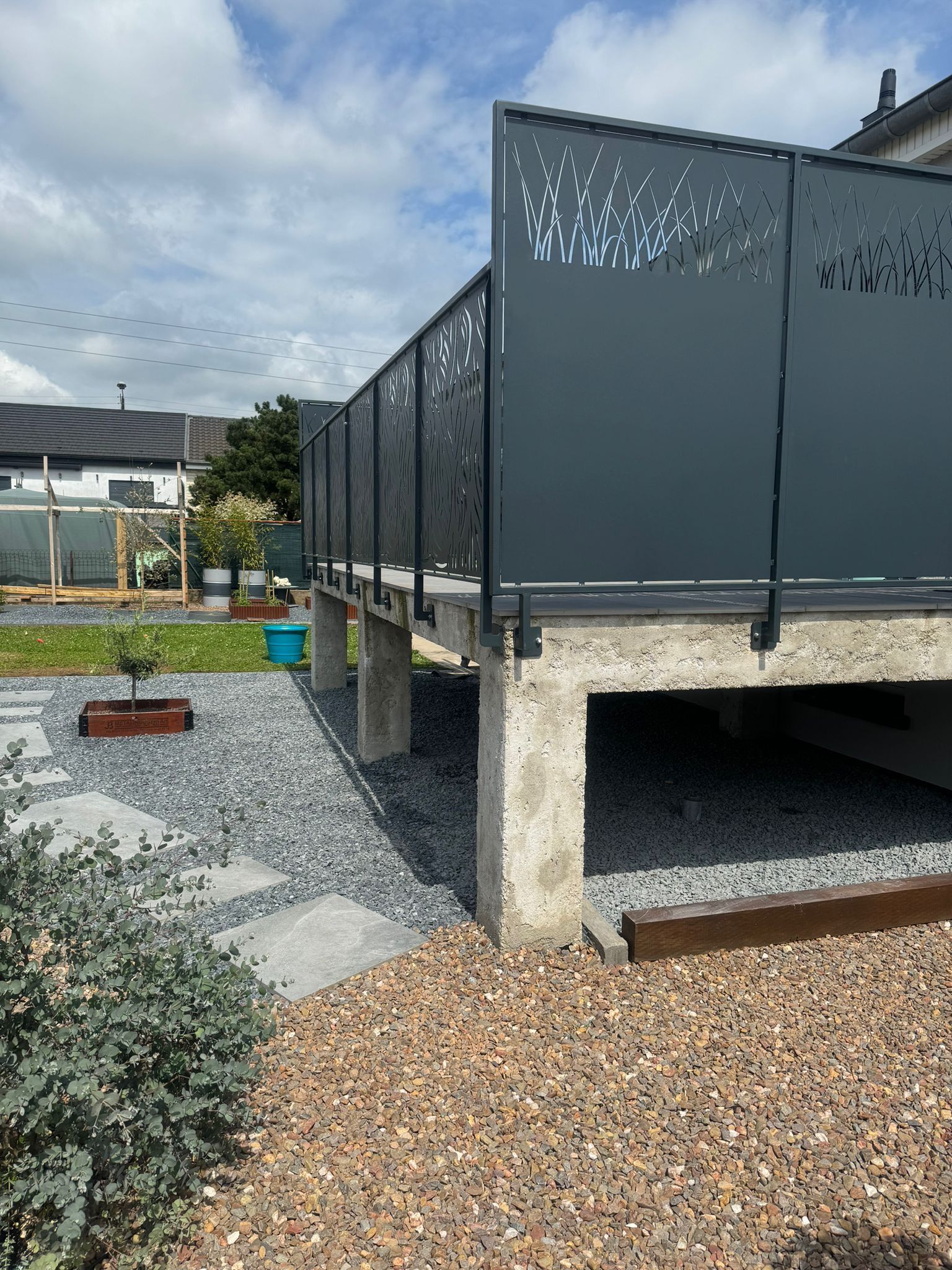 Terrasse grise avec rambarde métallique décorative sur piliers en béton, donnant sur une cour avec gravier et pavés.