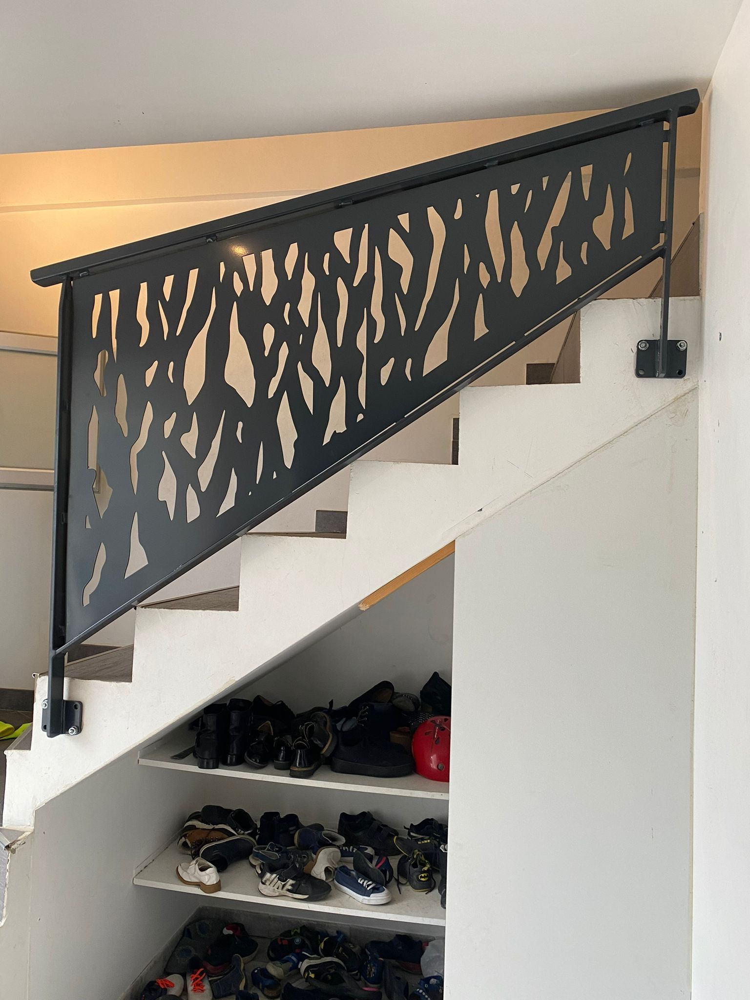 Rambarde en métal noire découpée au laser, ornée d'un motif organique, sur un escalier blanc. En dessous, un meuble à chaussures.