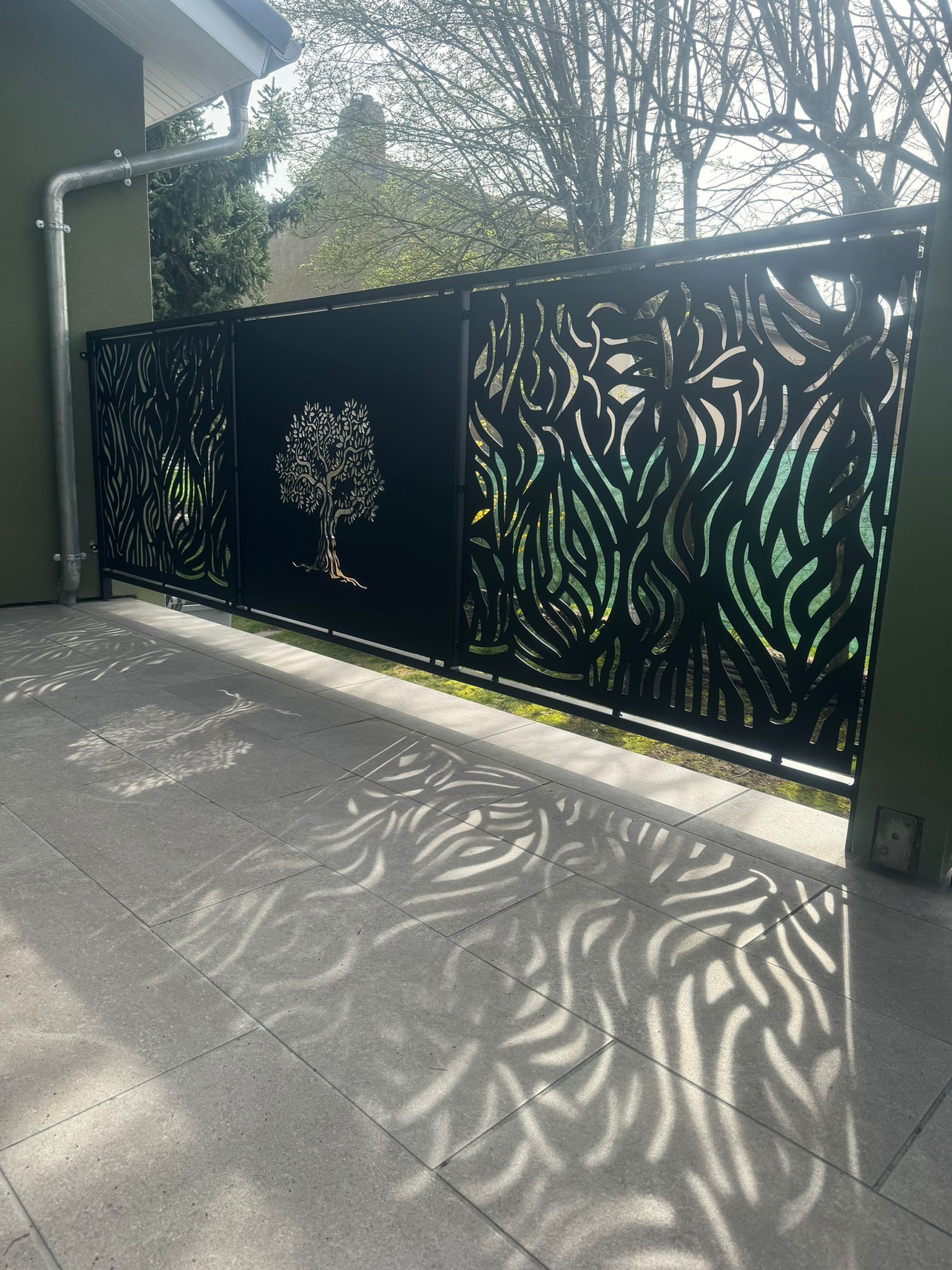 Des panneaux de clôture en métal noir découpés au laser, ornés de motifs inspirés de la nature projettent des ombres complexes sur une terrasse carrelée grise.