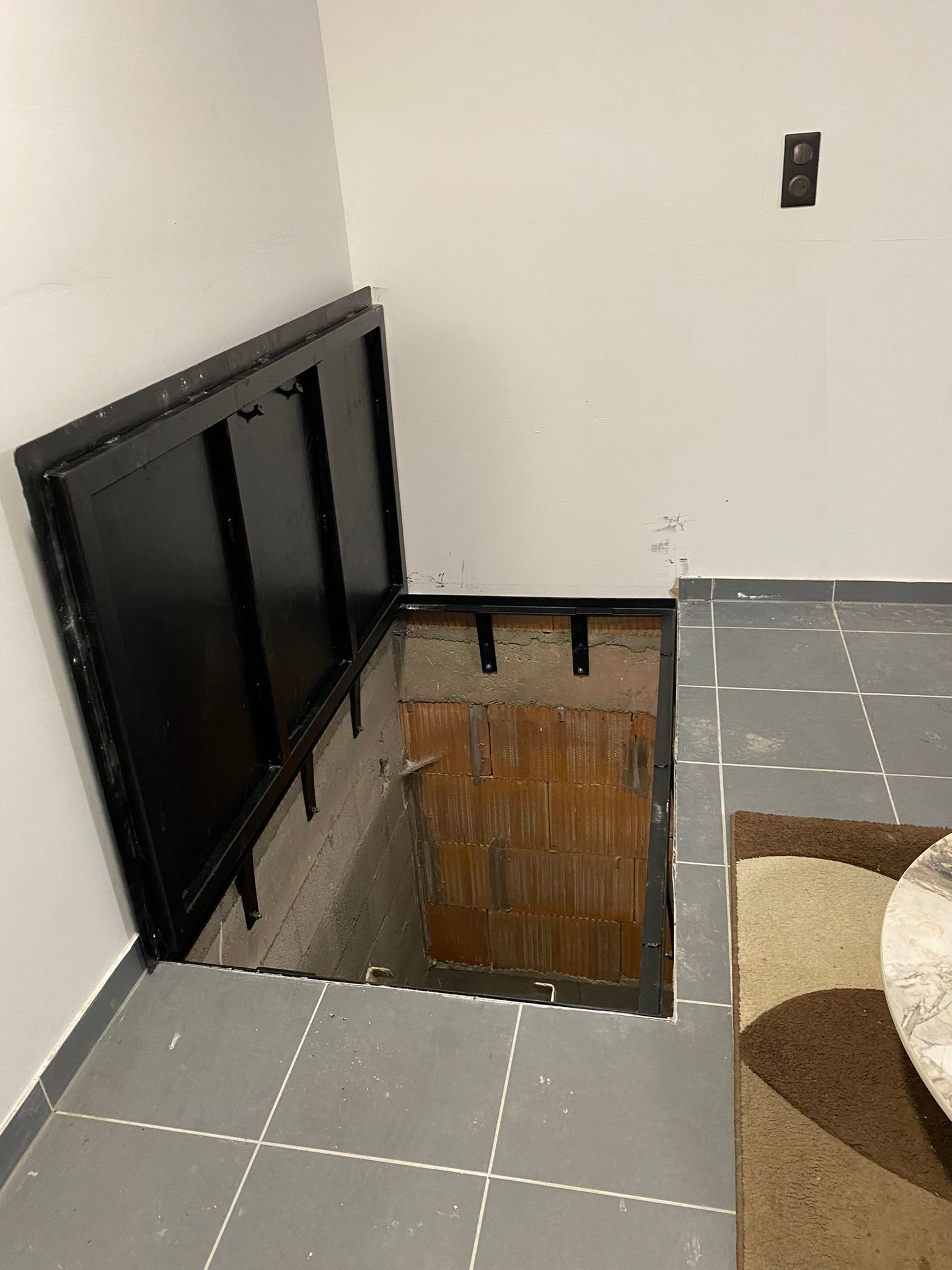 Trappe noire ouverte dans le sol carrelé, donnant accès à une cave aux murs de briques.