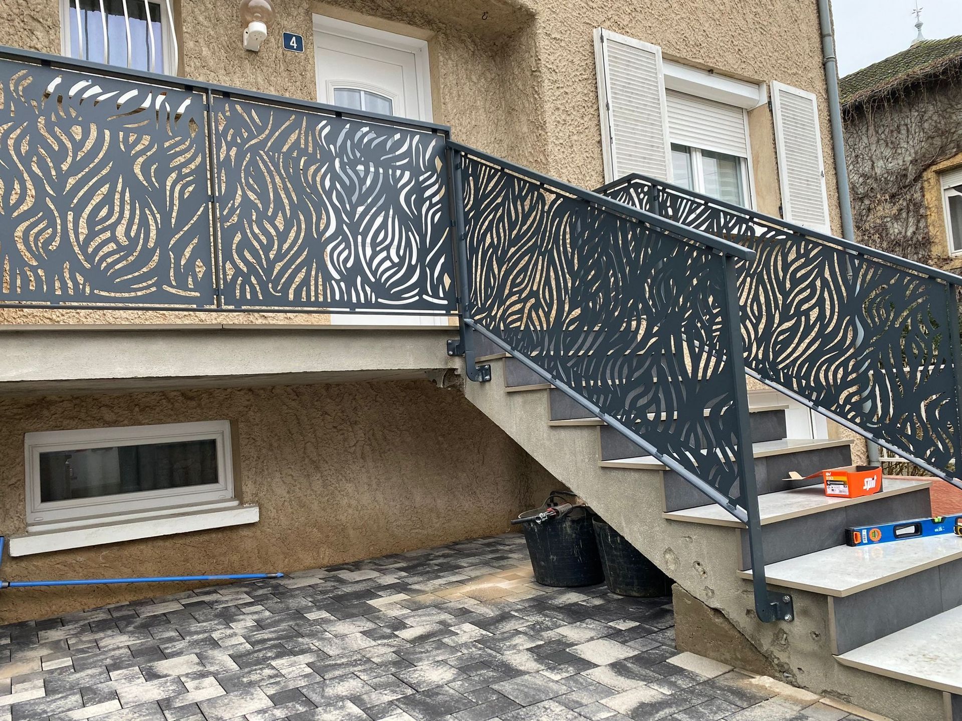 Escalier extérieur avec rampe en métal à motifs, gris, sur une maison avec terrasse en pierre.