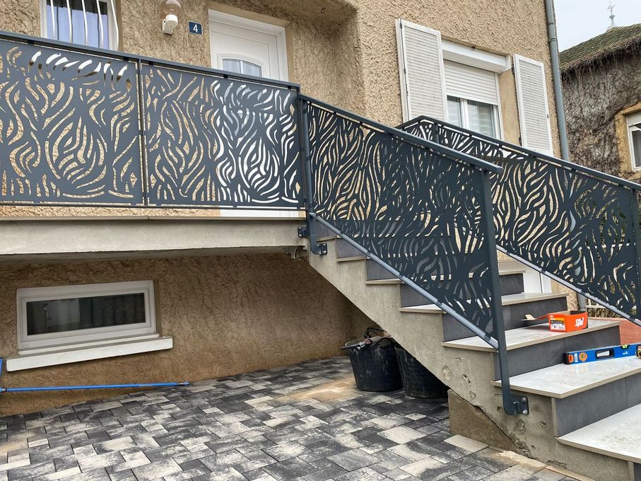 Escalier extérieur avec rampe en métal à motifs, gris, sur une maison avec terrasse en pierre.