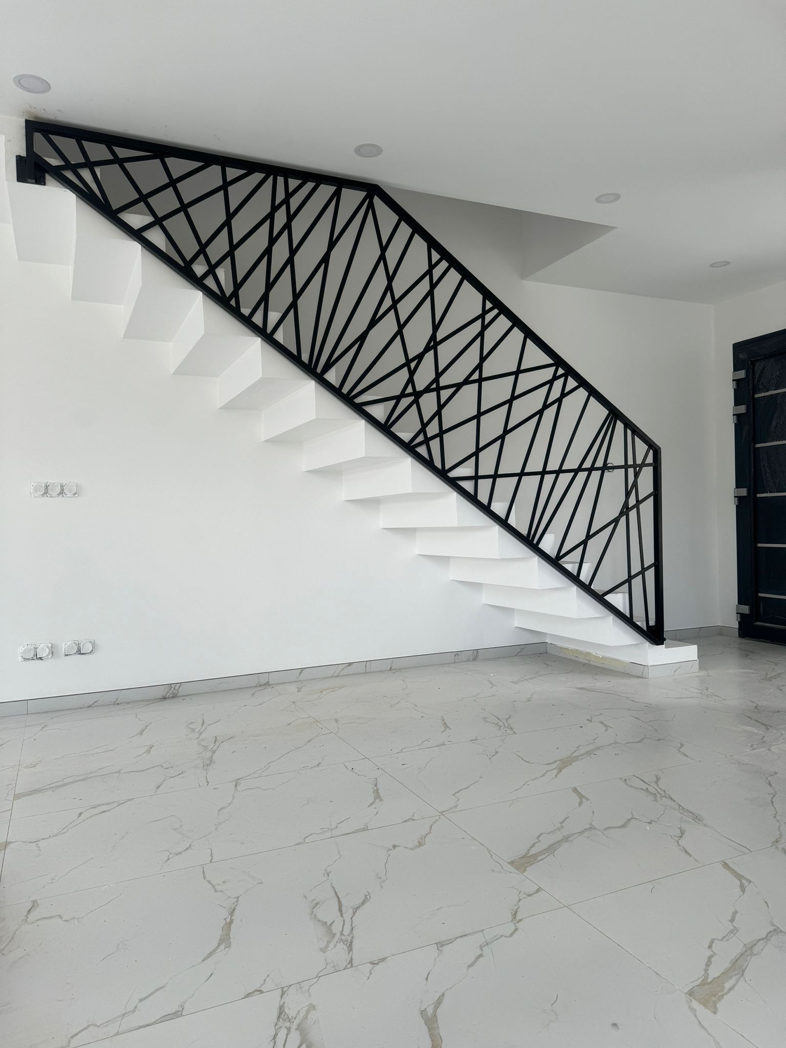 Escalier blanc avec rampe géométrique noire dans une pièce aux murs blancs et au sol en marbre.