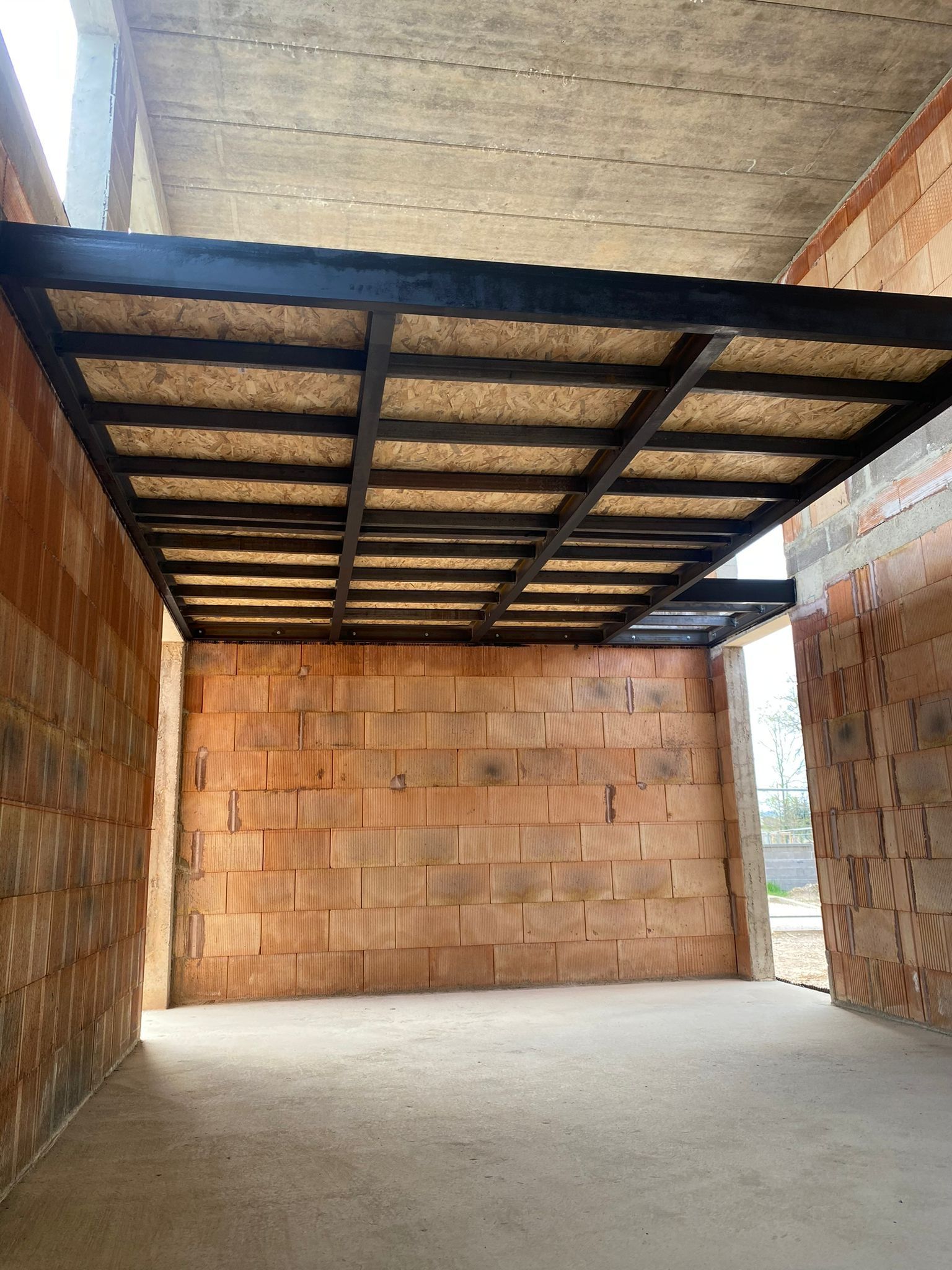 Une pièce en construction avec une structure métallique noire pour un deuxième étage. Les murs sont en briques rouges et le sol en béton.