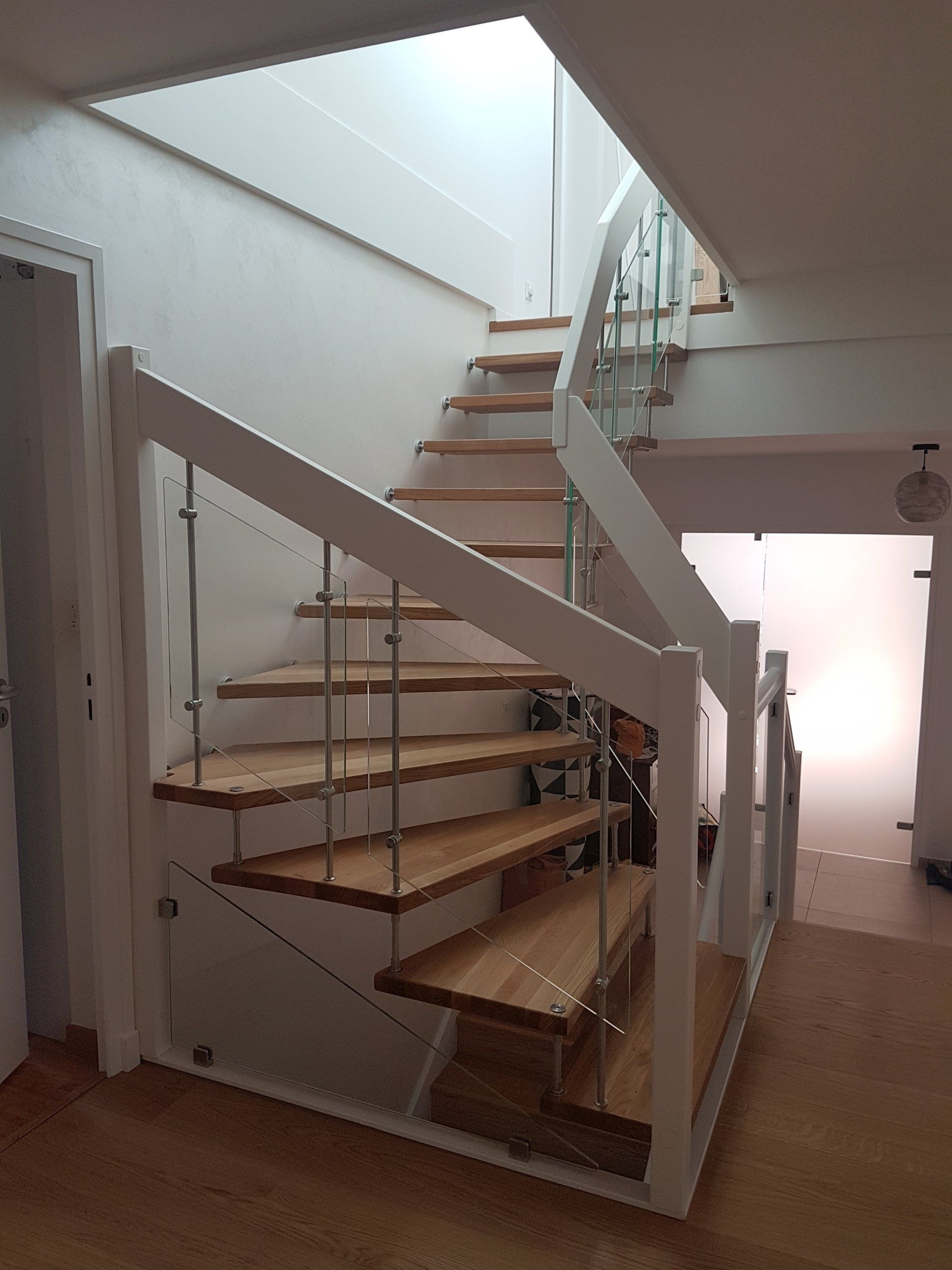 Escalier en bois avec rampes blanches et panneaux de verre, montant vers une lucarne.