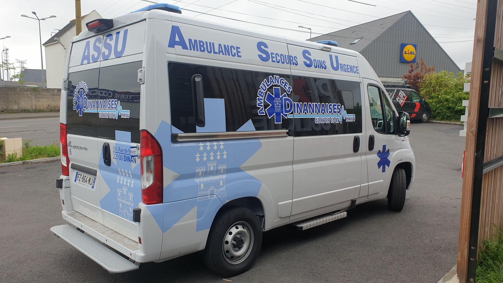 Ambulance blanche aux accents bleus, garée près d'un bâtiment. L'inscription Ambulance est visible.