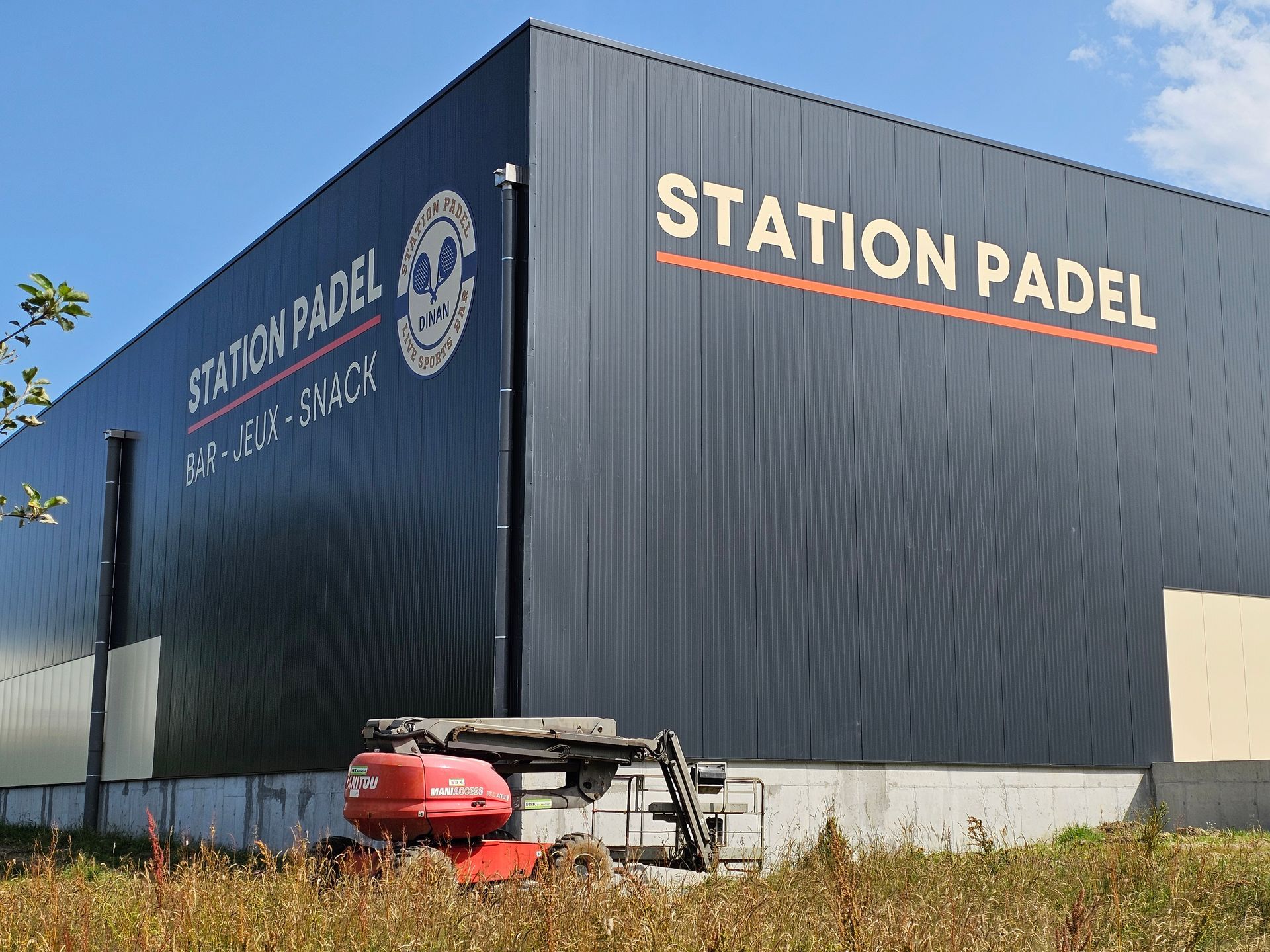 Bâtiment noir avec l'inscription STATION PADEL en lettres orange. Des engins de chantier sont stationnés devant le bâtiment.
