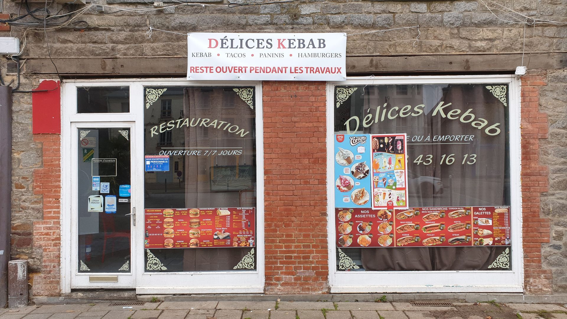Façade d'un restaurant de kebab nommé Délice Kebab avec vitrines présentant les plats du menu. Décoration en briques rouges.