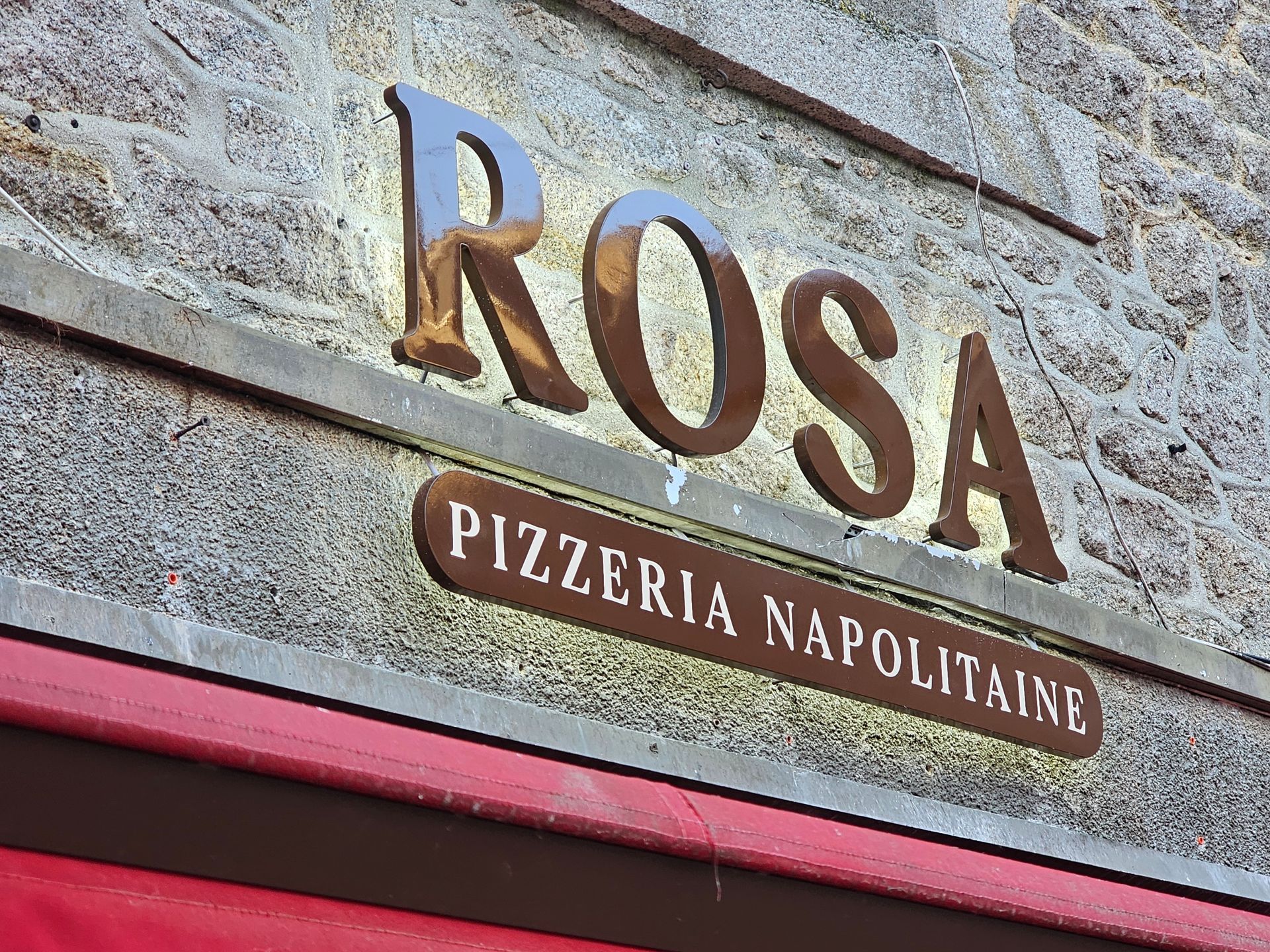 Panneau pour Rosa Pizzeria Napolitaine en lettres métalliques sur un mur de pierre, avec un auvent rouge en dessous.
