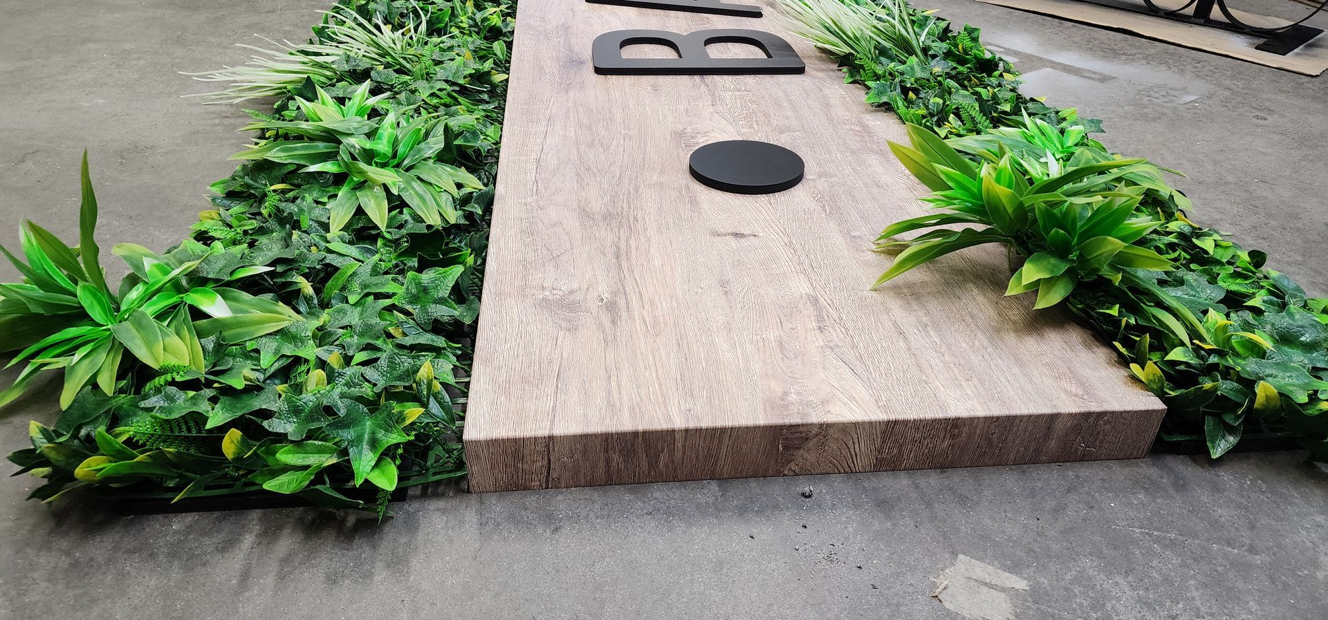 Un panneau en bois avec des lettres noires et un cercle, entouré de plantes vertes, sur une surface grise.