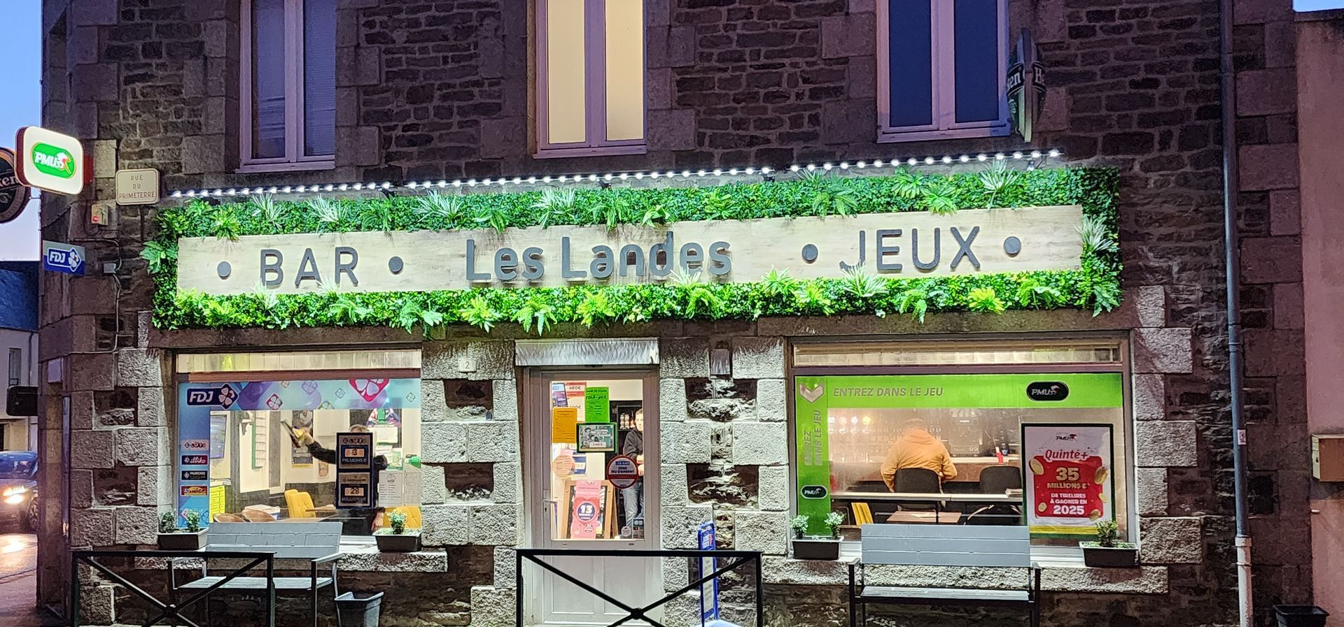 Un bâtiment en pierre avec une enseigne de bar : Les Landes Jeux. Extérieur avec fenêtres ouvertes et tables.
