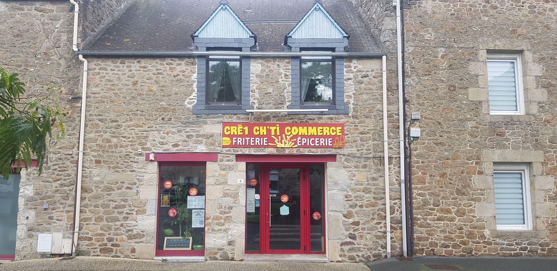 Un bâtiment en pierre avec un café aux cadres rouges. L'enseigne indique Café du Commerce. Deux petites fenêtres au-dessus.