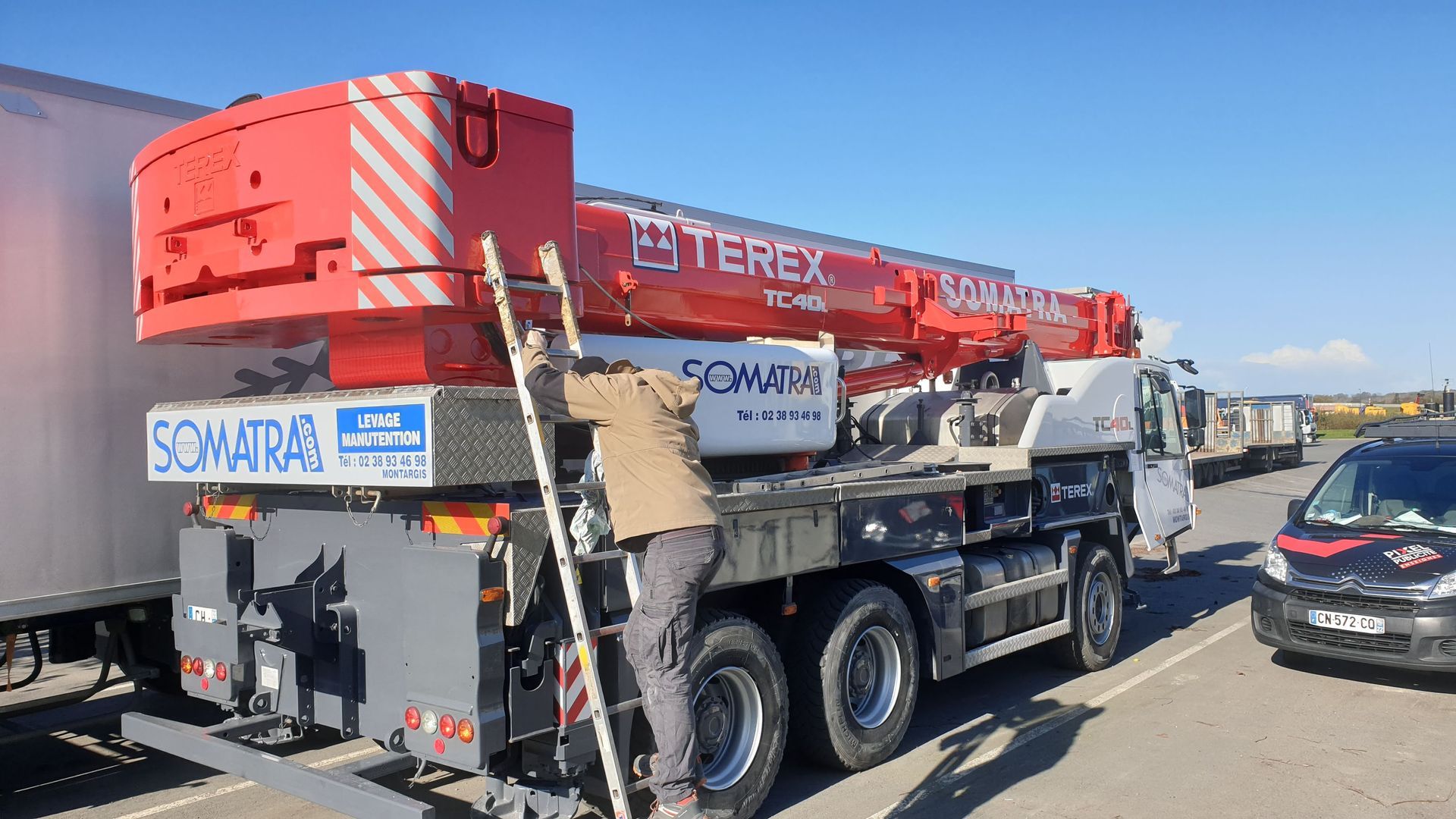 Une personne monte une échelle pour travailler sur une grue Terex rouge située sur un camion dans un parking.