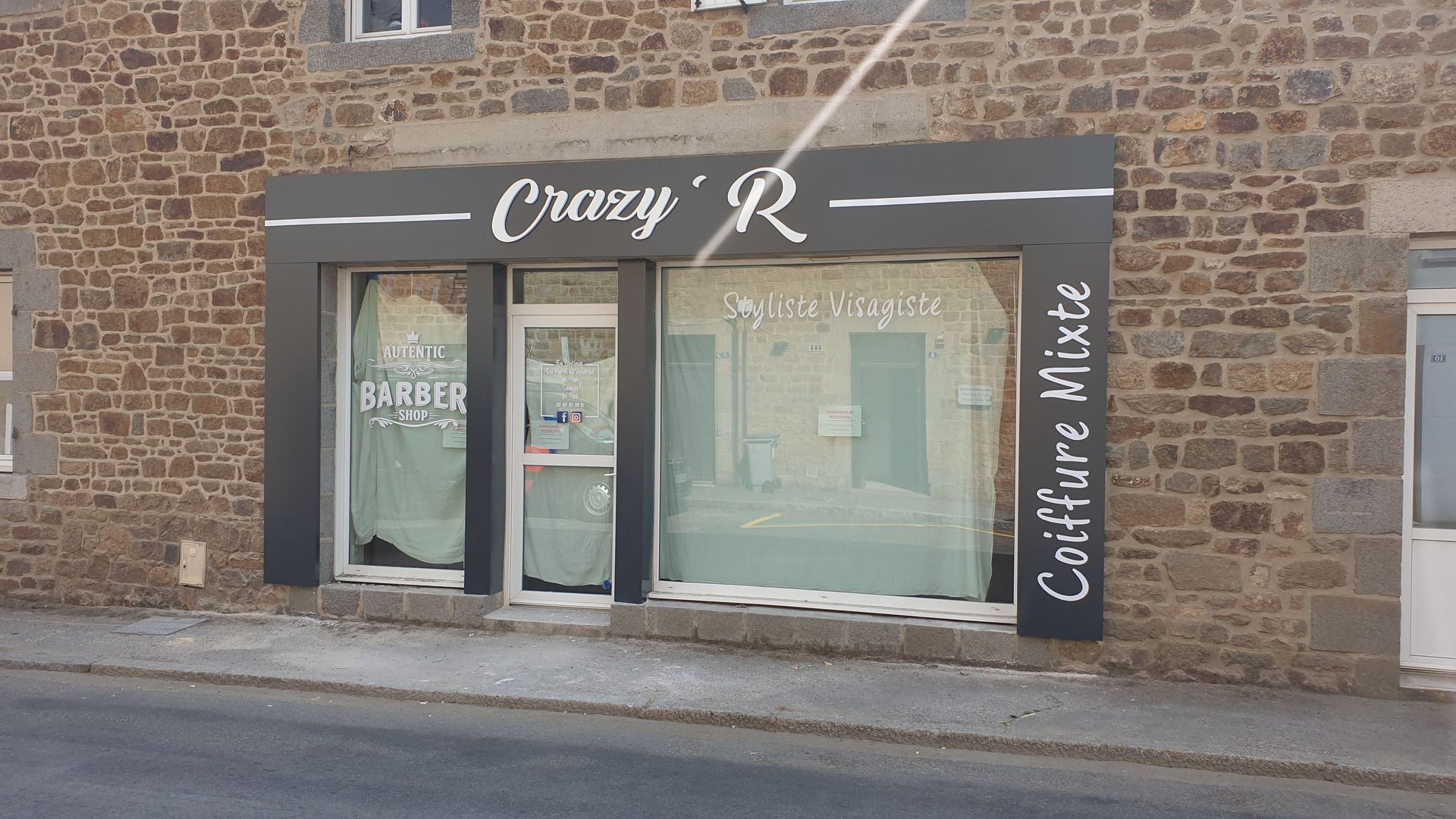 Extérieur du salon de coiffure Crazy' R dans un bâtiment en pierre. Cadre sombre autour des fenêtres et de la porte. Inscription sur le côté : Coiffure Mixte.