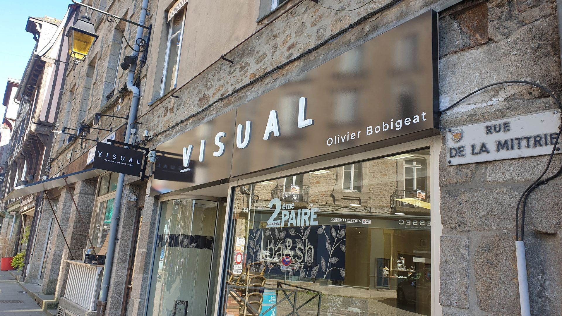 Une devanture de magasin étiquetée VISUAL en lettres blanches, avec une petite enseigne et un panneau de rue RUE DE LA MITRIE.