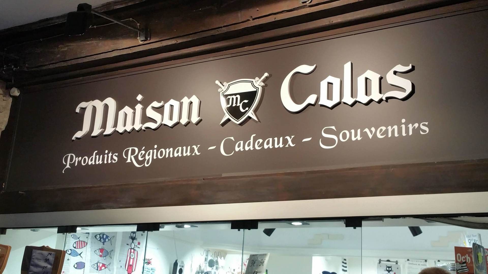 Enseigne de la Maison Colas, boutique de produits régionaux, cadeaux et souvenirs. Fond marron foncé avec lettrage blanc.