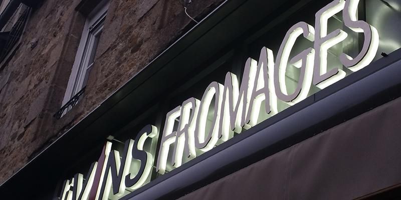 Panneau Vins Fromages en lettres blanches lumineuses au dessus d'une devanture de magasin la nuit.