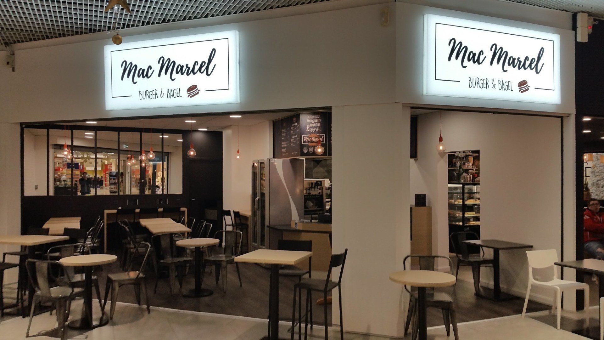 Extérieur du café Mac Marcel avec tables et chaises ; enseignes lumineuses ; décor blanc et noir.