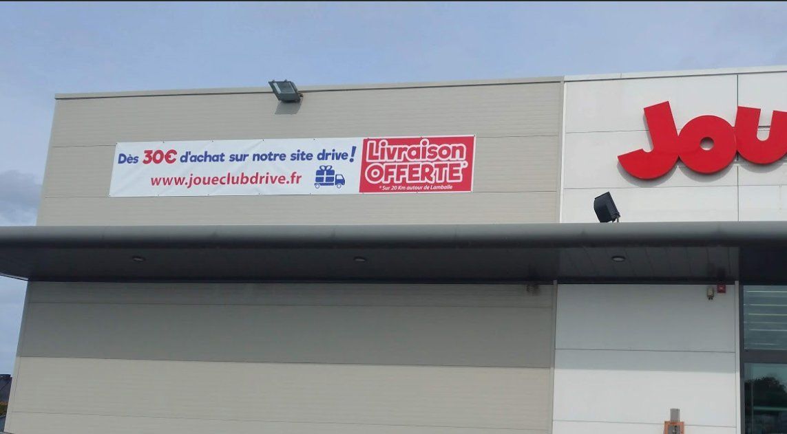 Devanture du magasin JouéClub avec enseigne Livraison offerte  et site web. Bâtiment gris et blanc, ciel couvert.