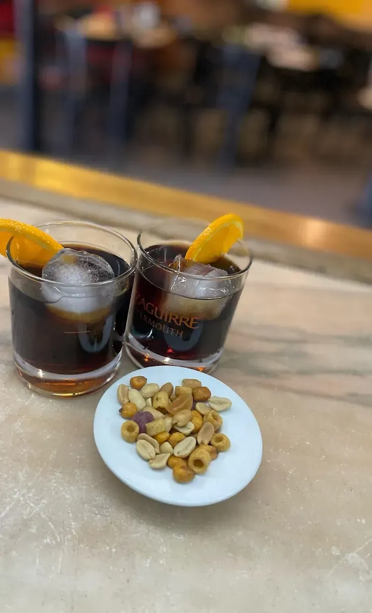 Dos cócteles oscuros con cáscaras de naranja y hielo, servidos con maní en una encimera de mármol.