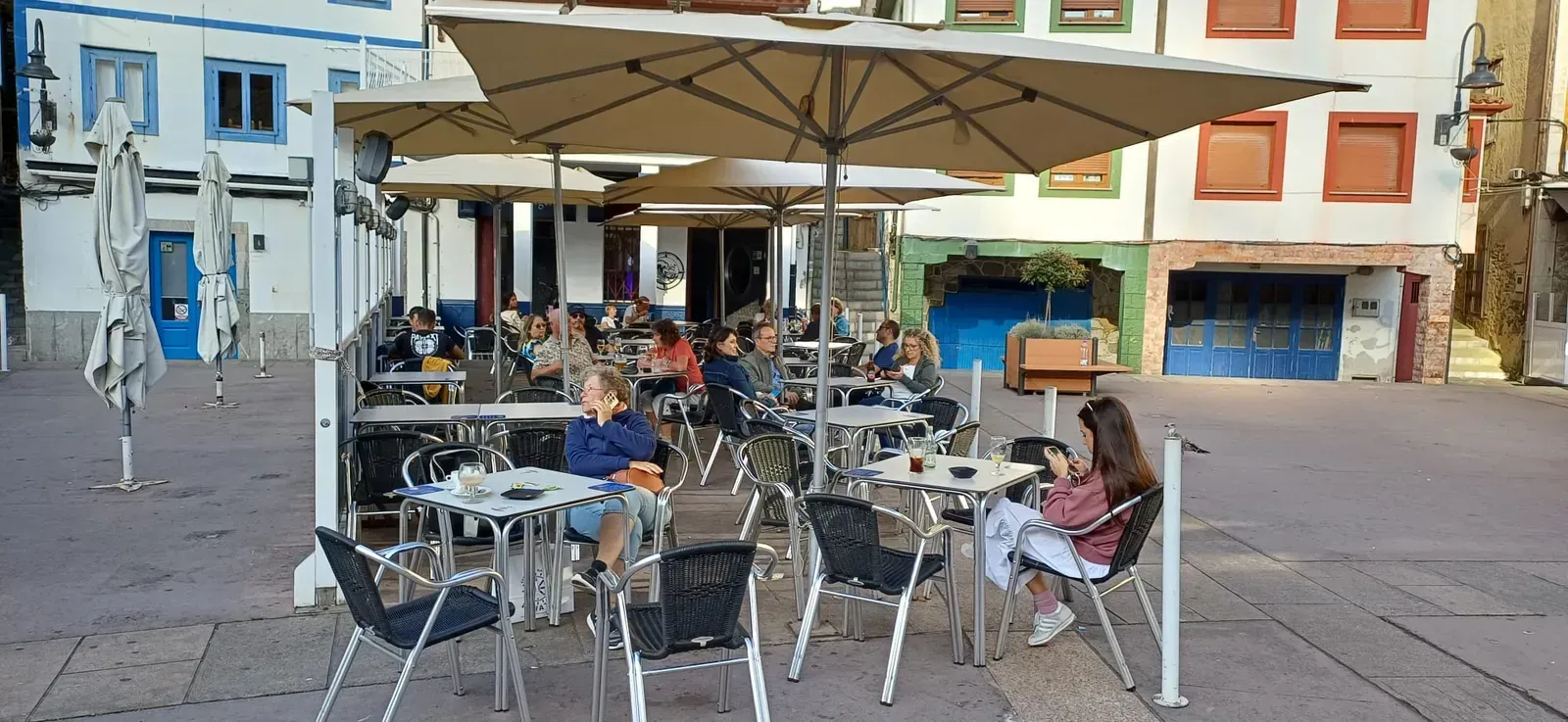 Edificios coloridos bordean una plaza con mesas y sombrillas; una ladera se eleva al fondo.