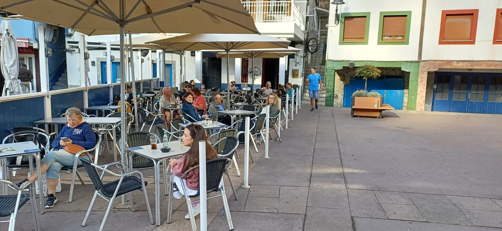 Edificios coloridos bordean una plaza con mesas y sombrillas; una ladera se eleva al fondo.