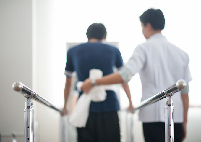 Un homme en chemise bleue marche avec l'aide d'un professionnel de la santé le long de barres parallèles.