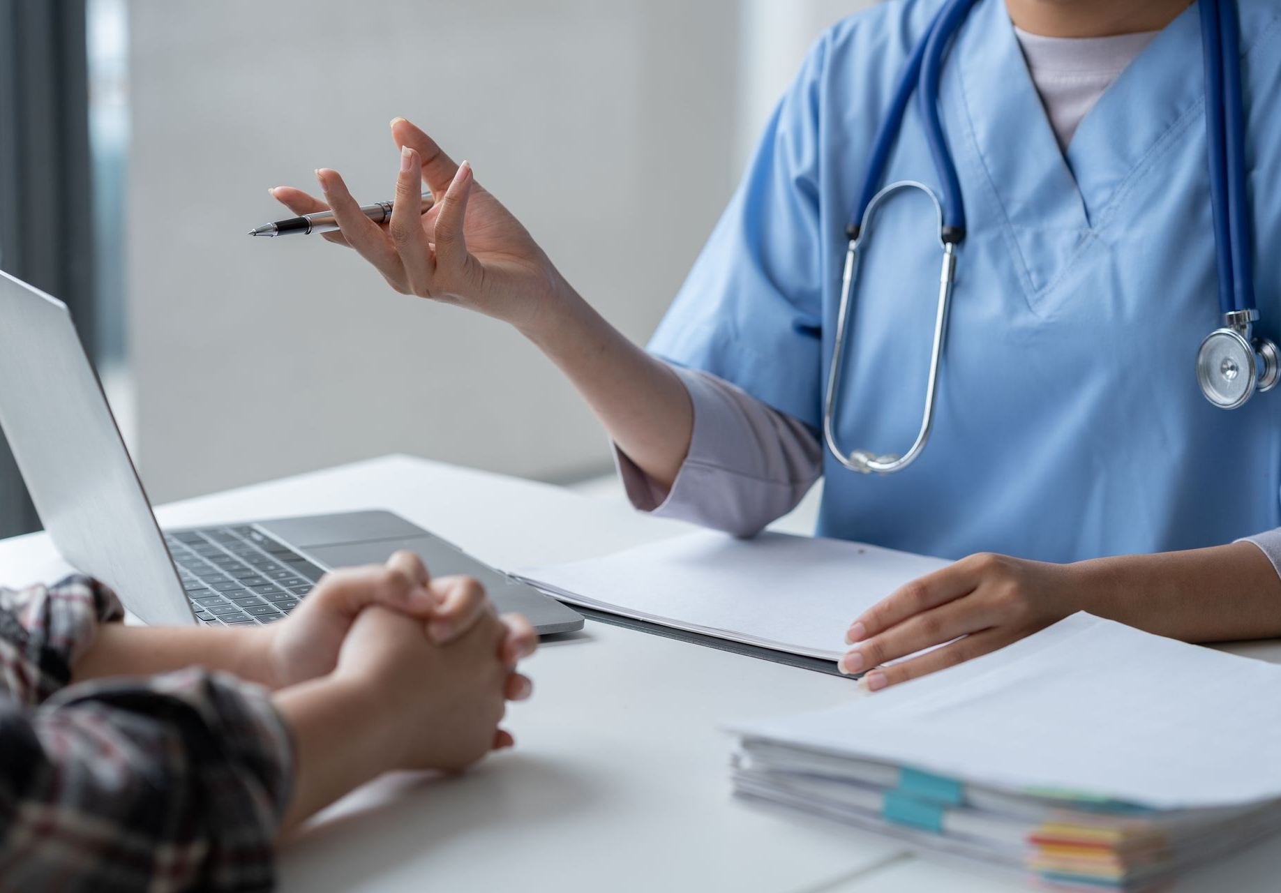 Un médecin en blouse bleue consulte un patient à un bureau, un ordinateur portable et des documents à portée de main.