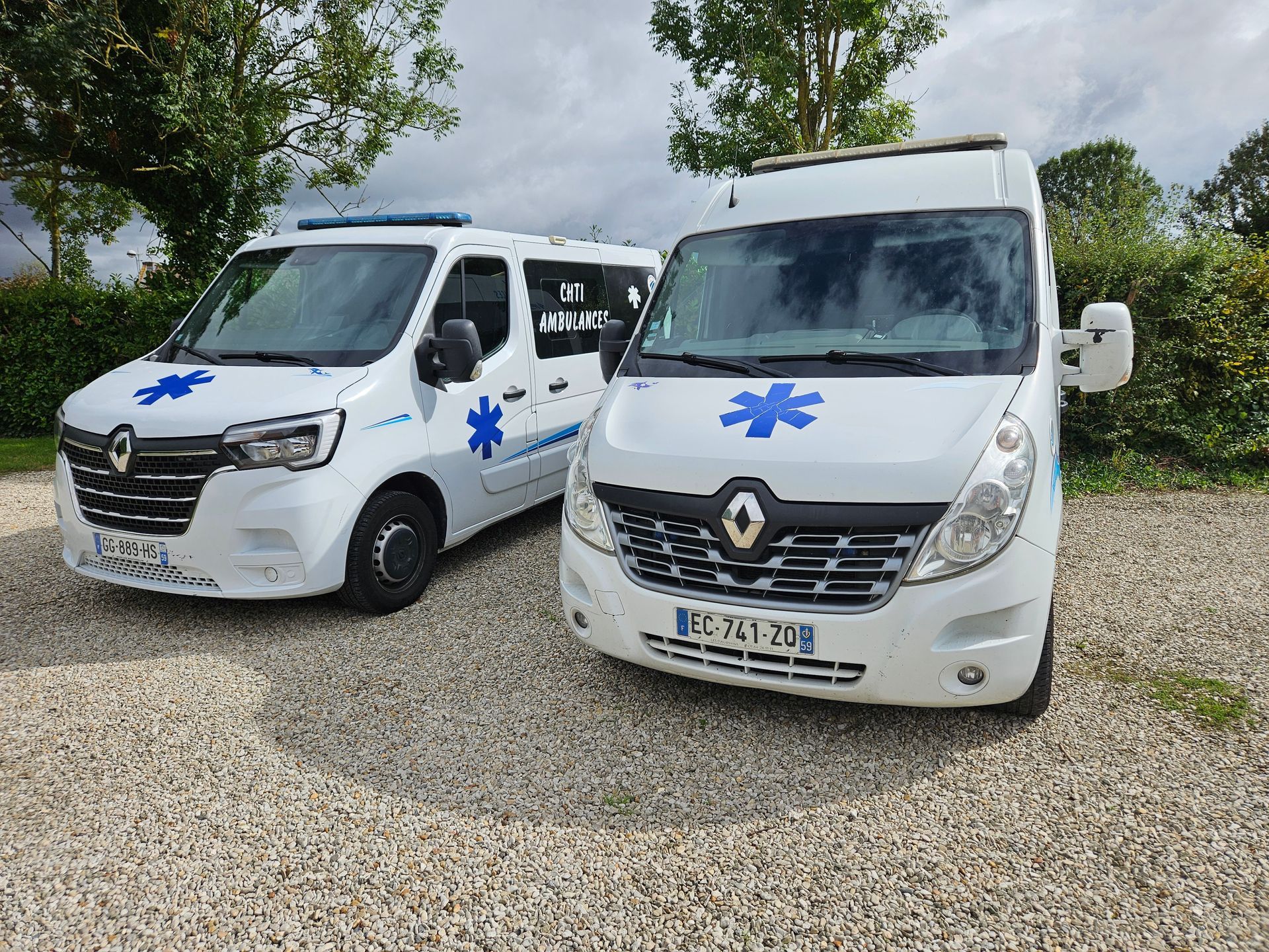 Deux ambulances blanches ornées d'étoiles bleues de vie sont stationnées sur du gravier sous un ciel nuageux.