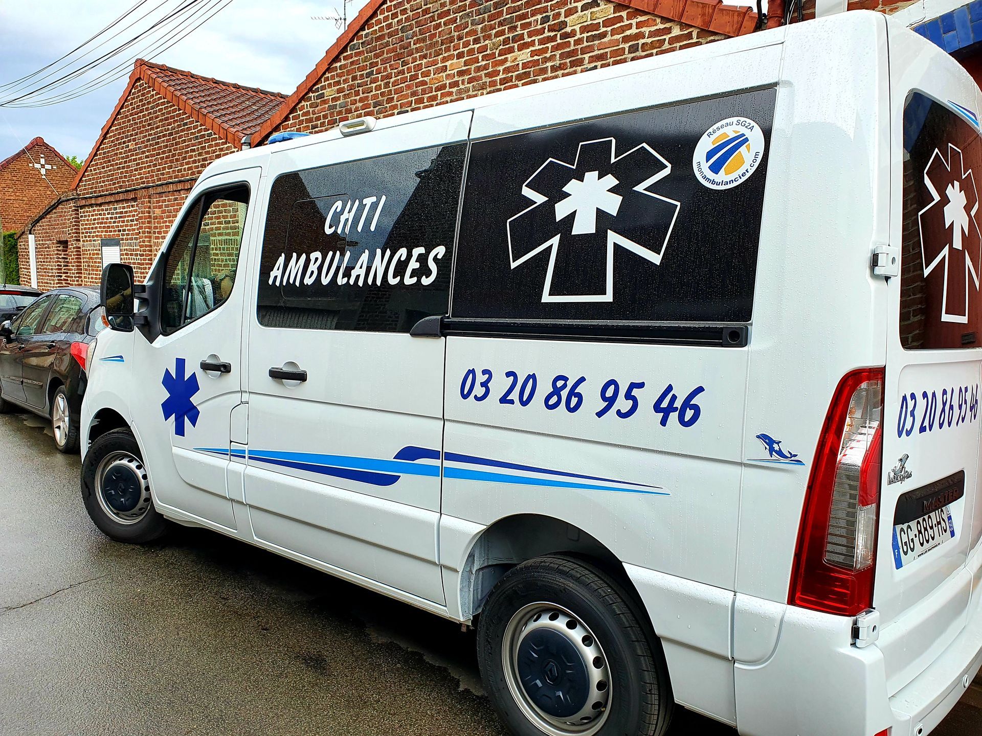 Ambulance blanche avec des accents bleus, CH'TI AMBULANCES, stationnée dans une rue. Numéro de téléphone visible.