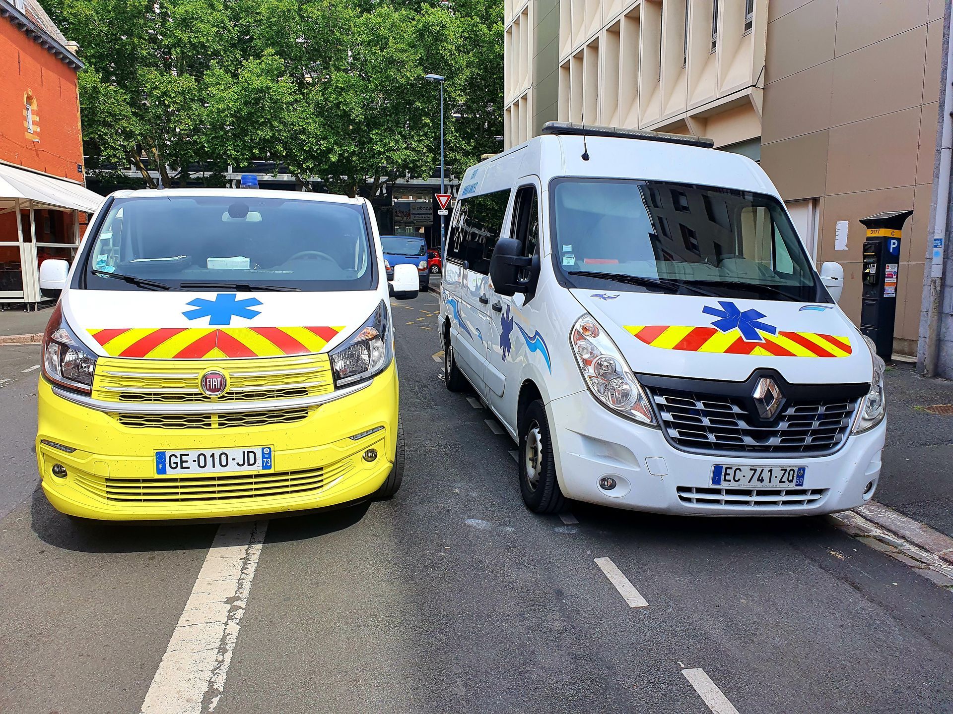 Deux ambulances sont stationnées dans une rue. L'une est jaune, l'autre blanche, toutes deux équipées de gyrophares et arborant l'étoile de vie.
