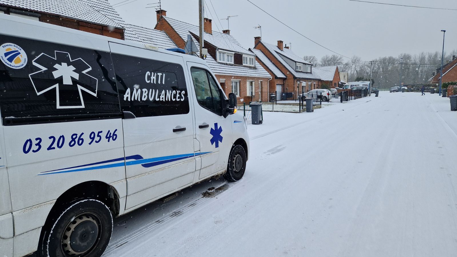 Une ambulance blanche avec des touches de bleu est garée dans une rue enneigée, devant des maisons en briques.