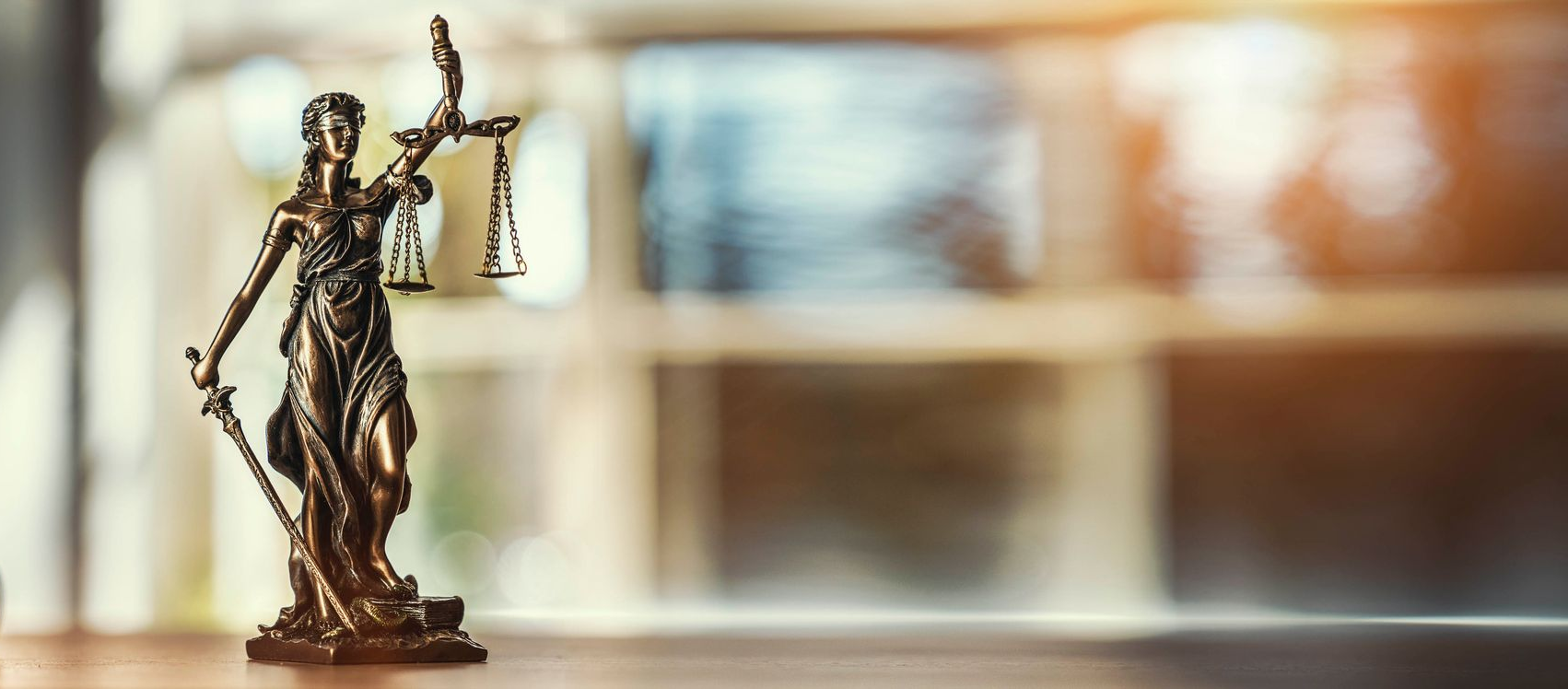 Bronzestatue der Justitia mit Waage und Schwert vor verschwommenem Hintergrund.