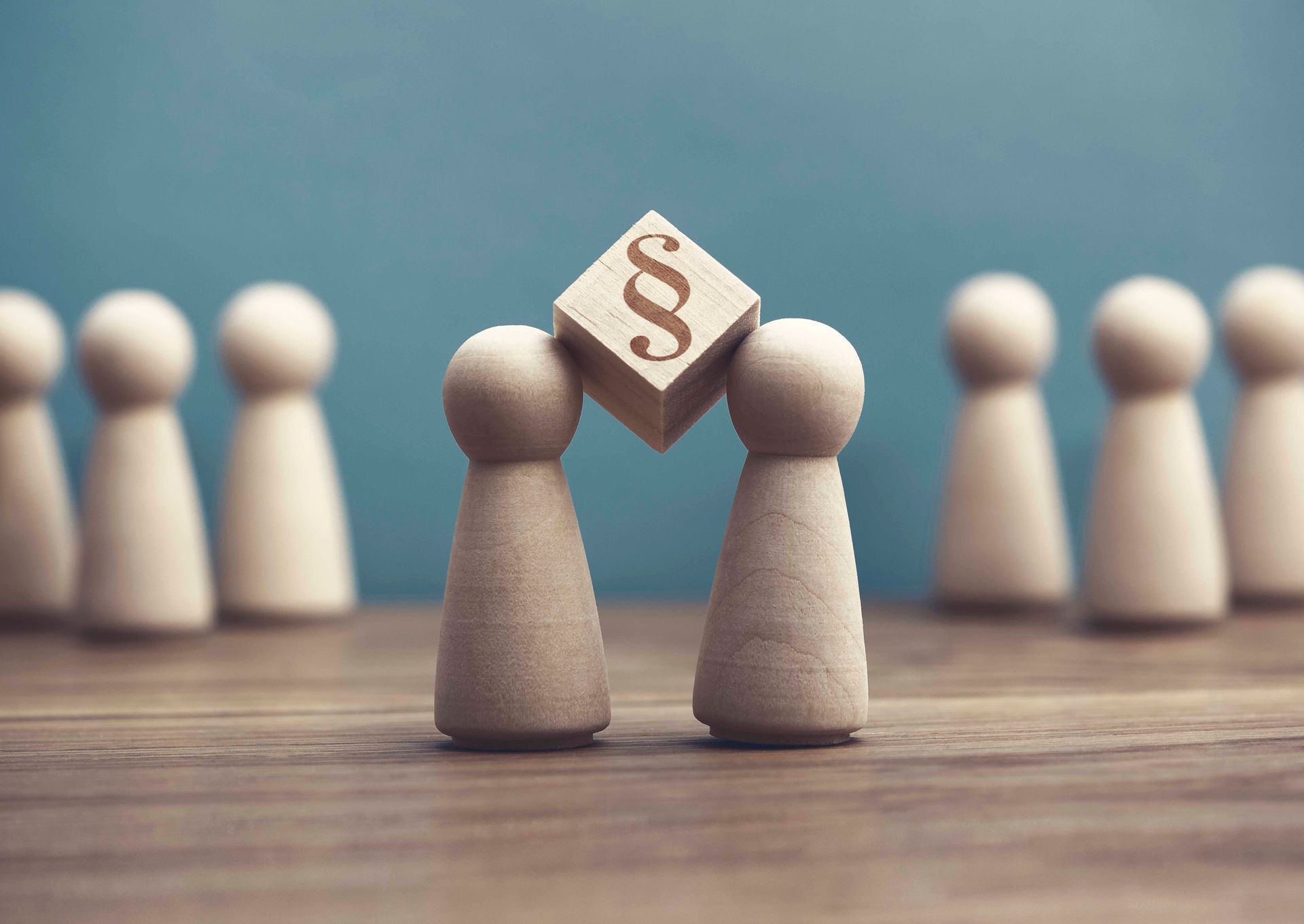 Zwei Holzfiguren halten einen Block mit einem Absatzsymbol; weitere Figuren im Hintergrund.