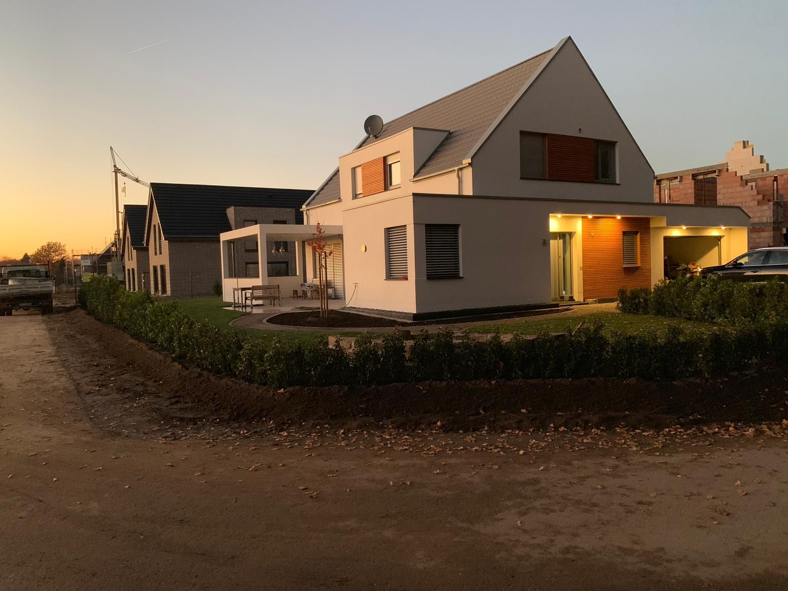 Modernes weißes Haus mit Giebeldach und Gartenanlage vor dem Hintergrund eines Sonnenuntergangs.